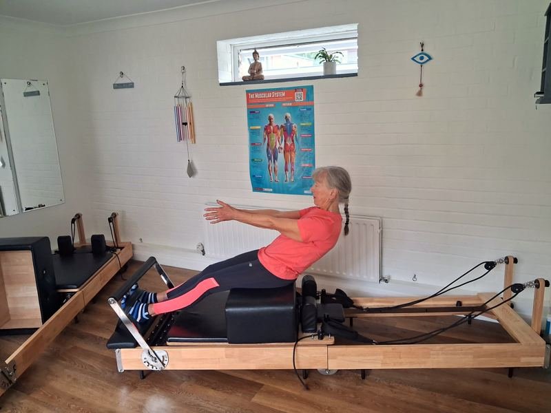 A client sitting on the reformer box, leaning back with arms reaching forward